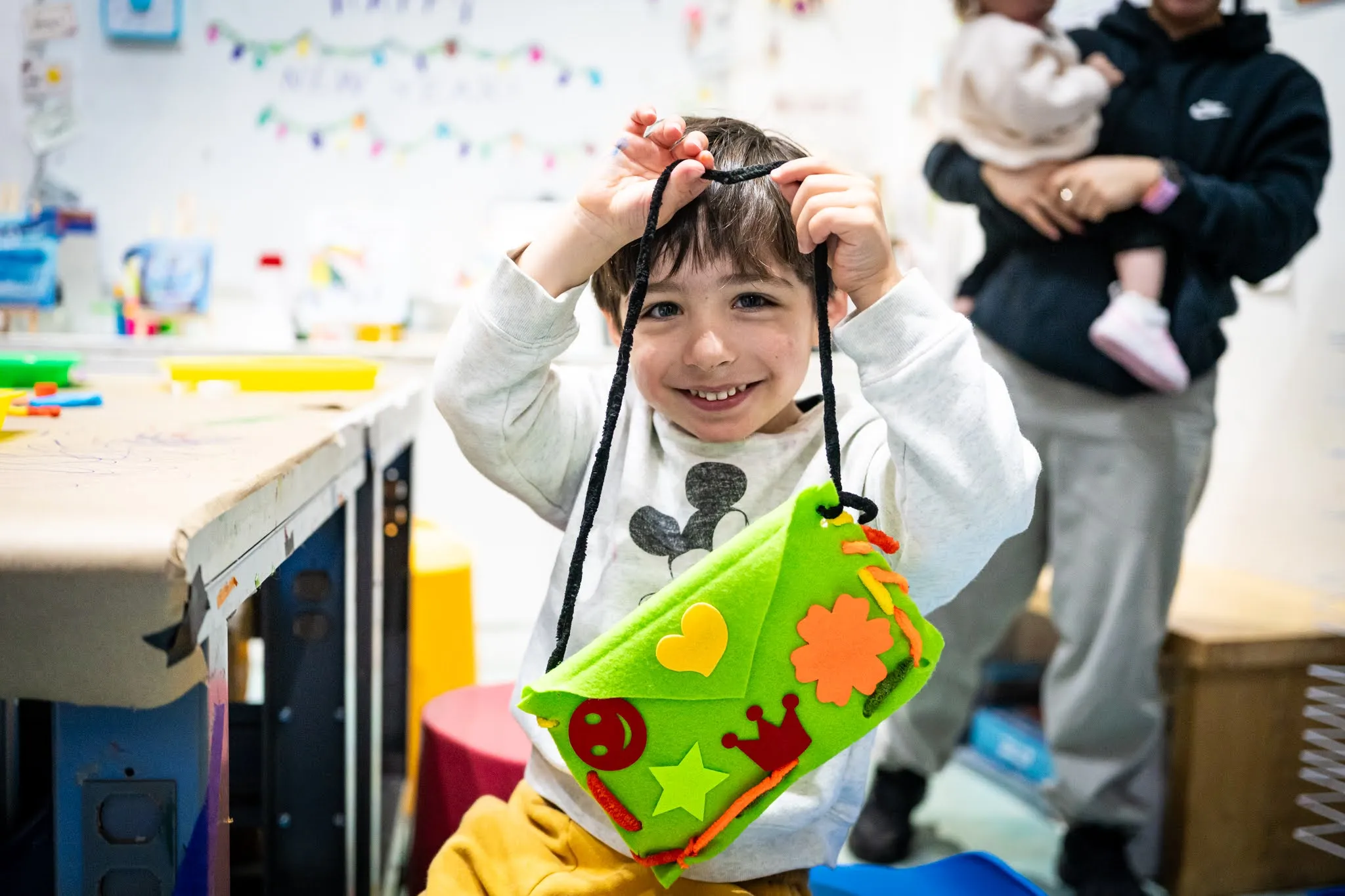 A smiling child holds up what appears to be a crafted felt purse or pouch that she has made. He is smiling at the camera.