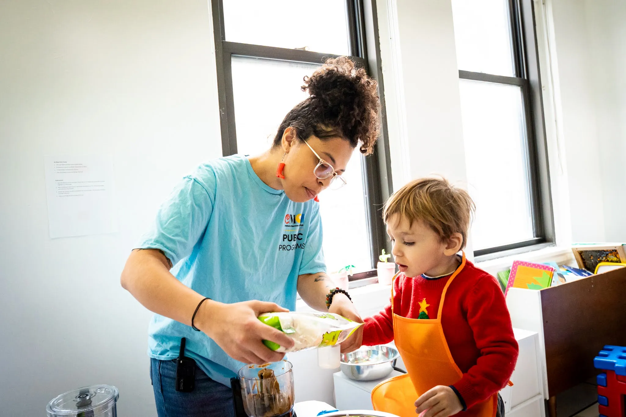 A young adult in a "Children's Museum of Manhattan Public Programs" shirt measures out something from a small plastic bag into a bowl beneath the bag. A child watches her carefully.