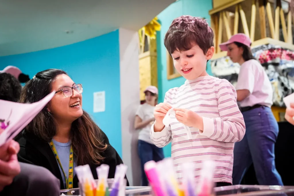 An adult woman smiles at a child, who appears to be inspecting some paper item or craft that he holds in his hands. In the background are more young adults wearing baseball caps.