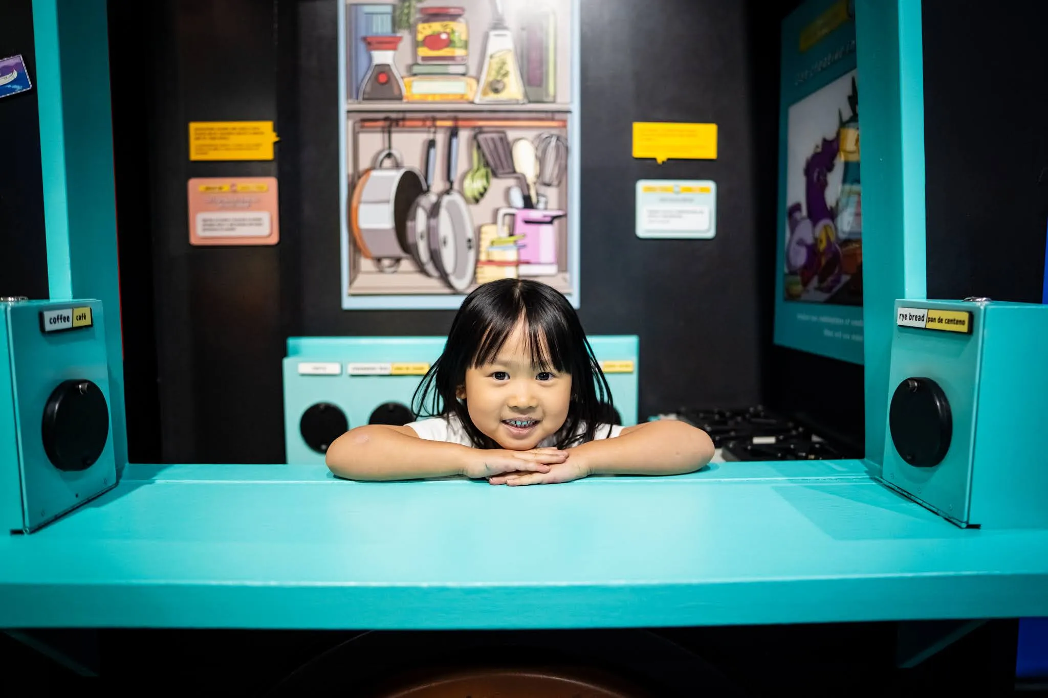 A child rests her hands on a table and her chin on top of her hands. The table is turquoise and flanked by boxes that look as if they may include or be speakers.