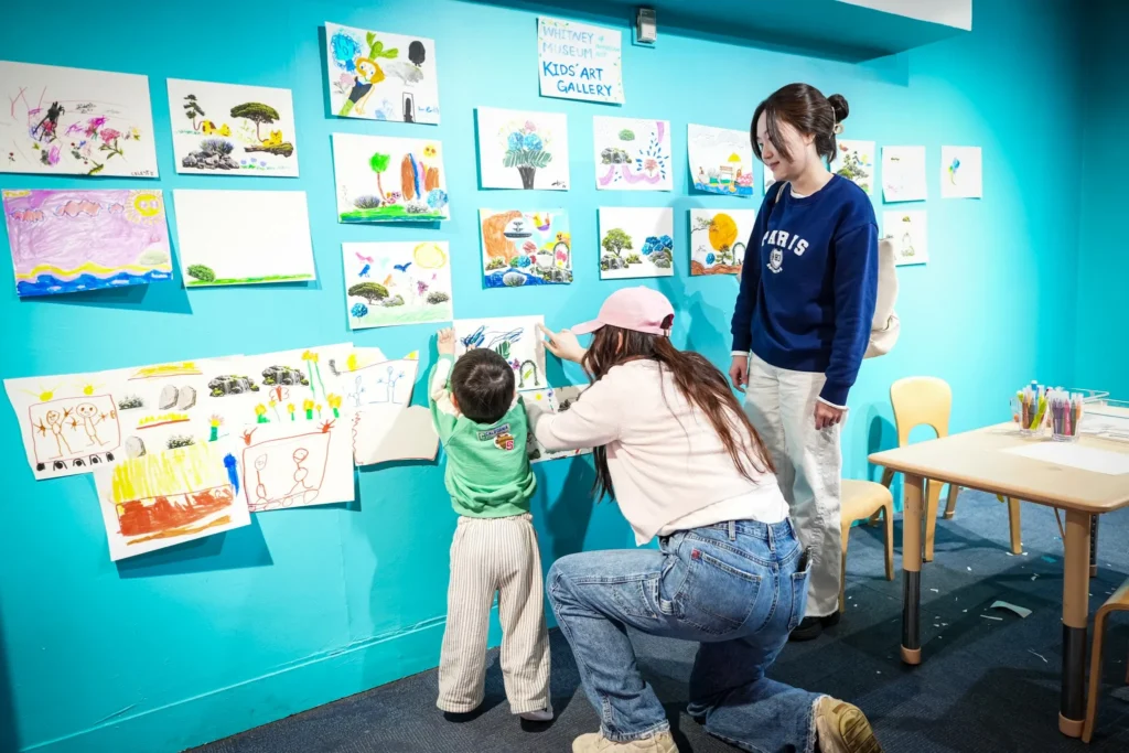 A bright blue wall has a sign that says "Whitney Museum Kids' Art Gallery" above a sample of over a dozen children's drawings. A child and adult have their backs to the camera while they adhere an additional drawing to the wall. A second adult looks on from profile.