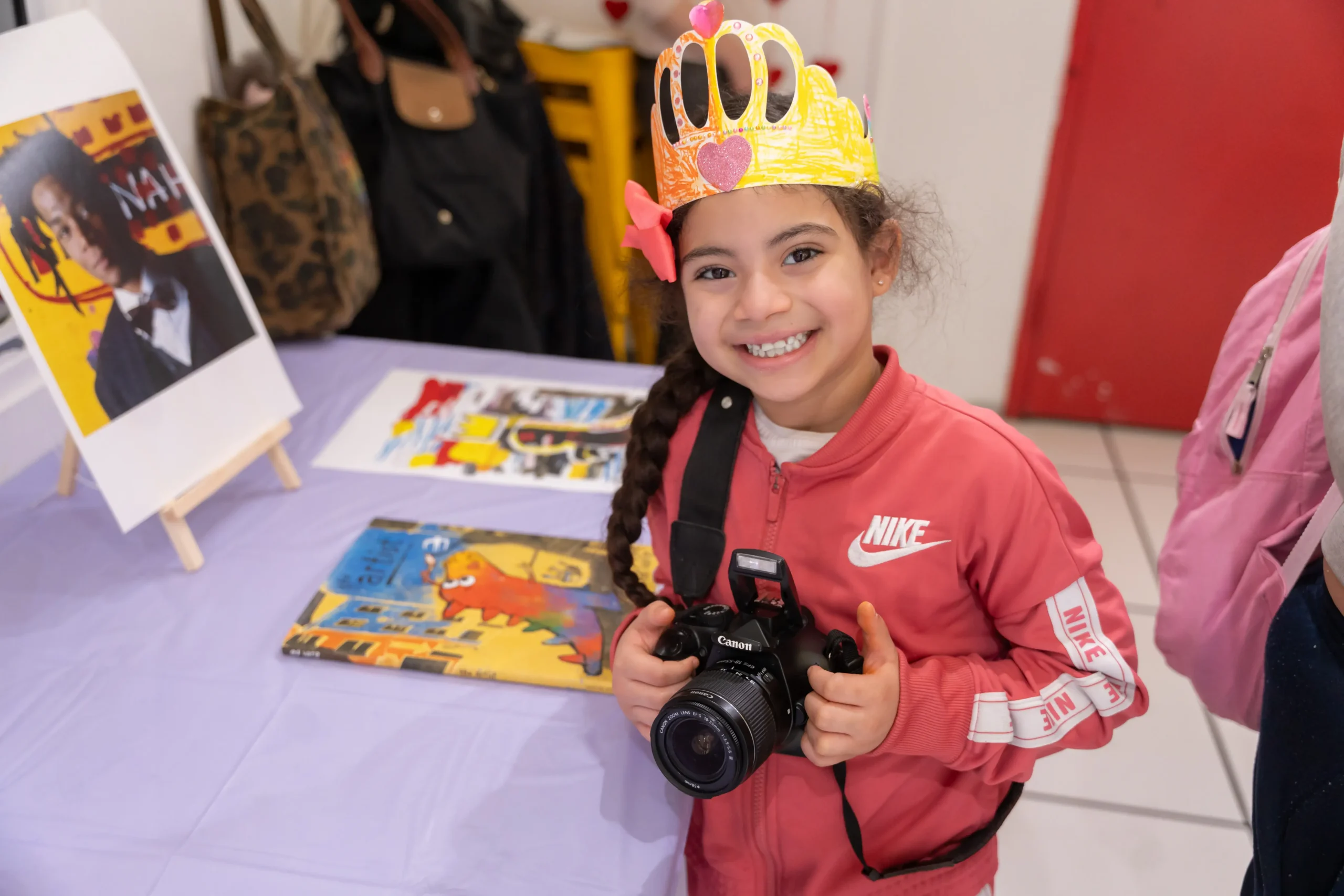 A child wears a paper crown and holds a large Canon camera while she smiles at this camera. Beside her is a table two what appears to be two books, and a stand of a photo of a man.