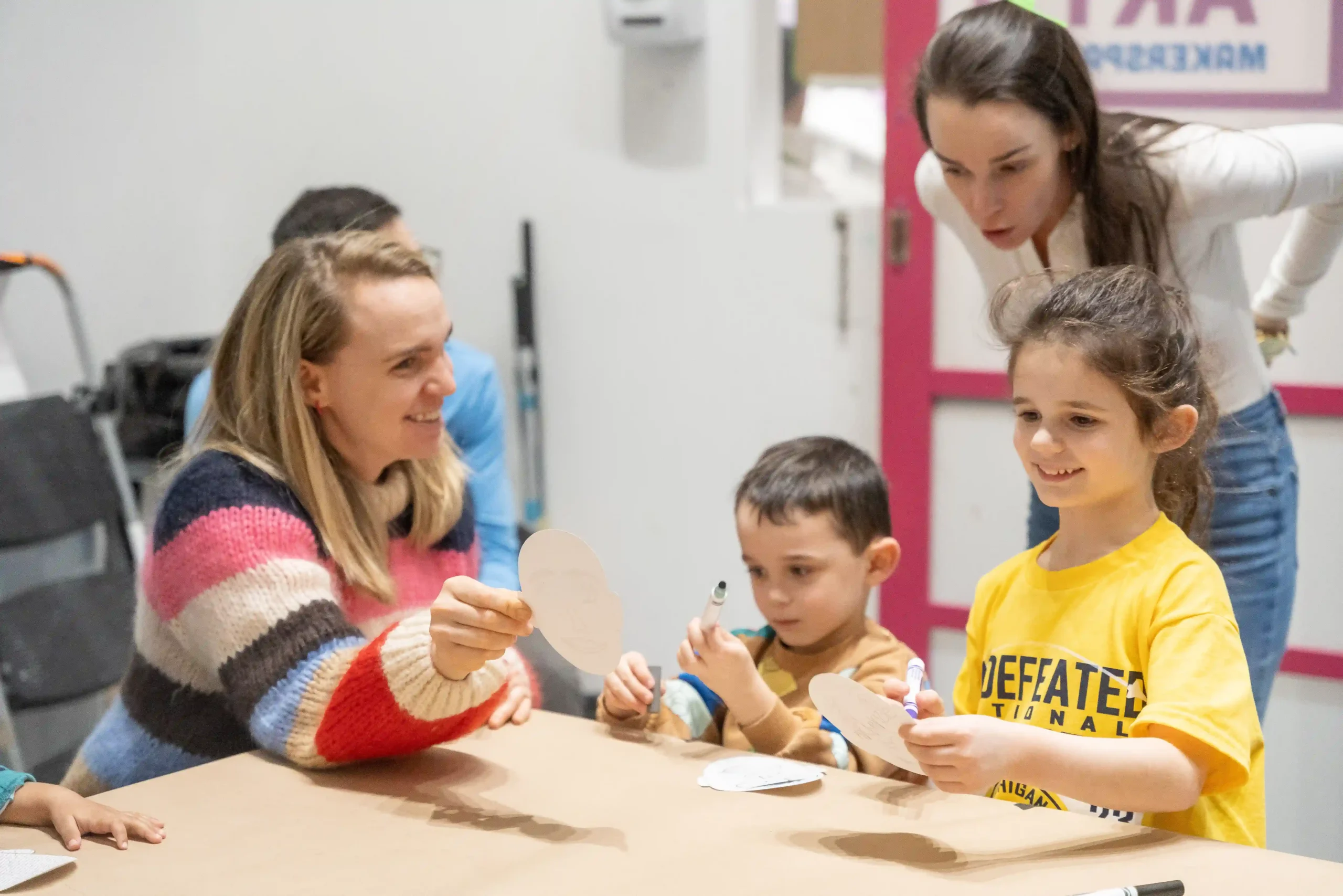 An adult smiles and holds up a paper circle, showing it to two small children and another adult who stands behind the children. The children both hold markers and one holds their own paper circle.