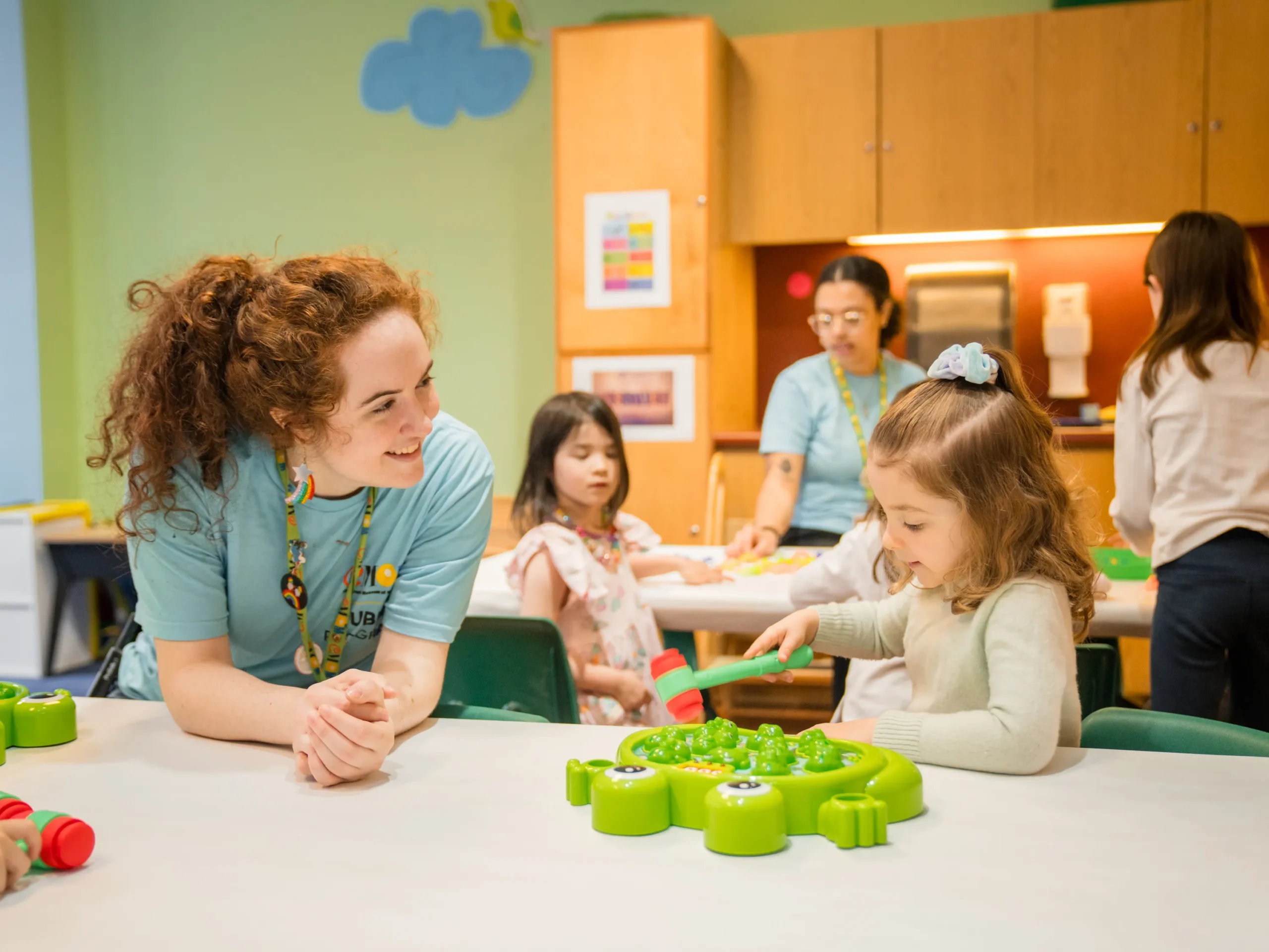 A young adult wears a "Children's Museum of Manhattan Public Programs" and leans down onto a table next to a child, who holds a mallet that may be part of a plastic frog game, which is on the table in front of her. Behind the two is another table with at least one child and one additional Public Programs staff member.