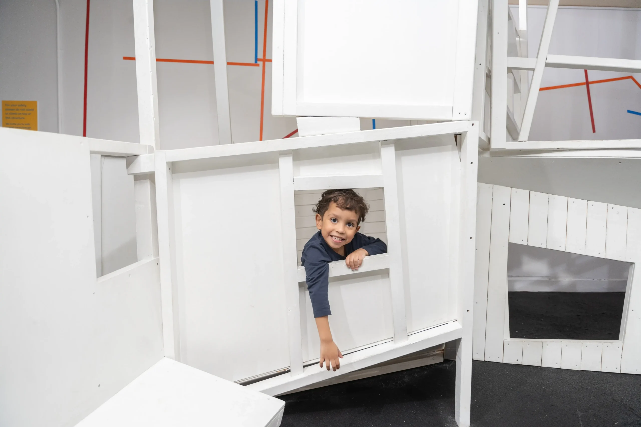 A child is smiling through the window of an all-white indoor playspace.