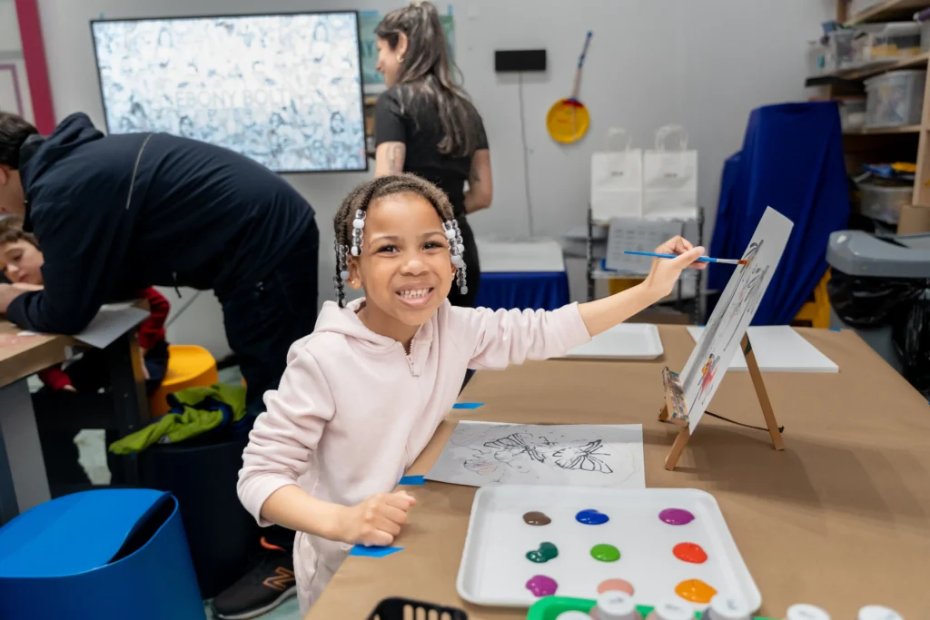 A child has a paint brush to a small canvas on a stand on a table. Next to the child is a palette with different colors of paints. The child smiles at the camera.