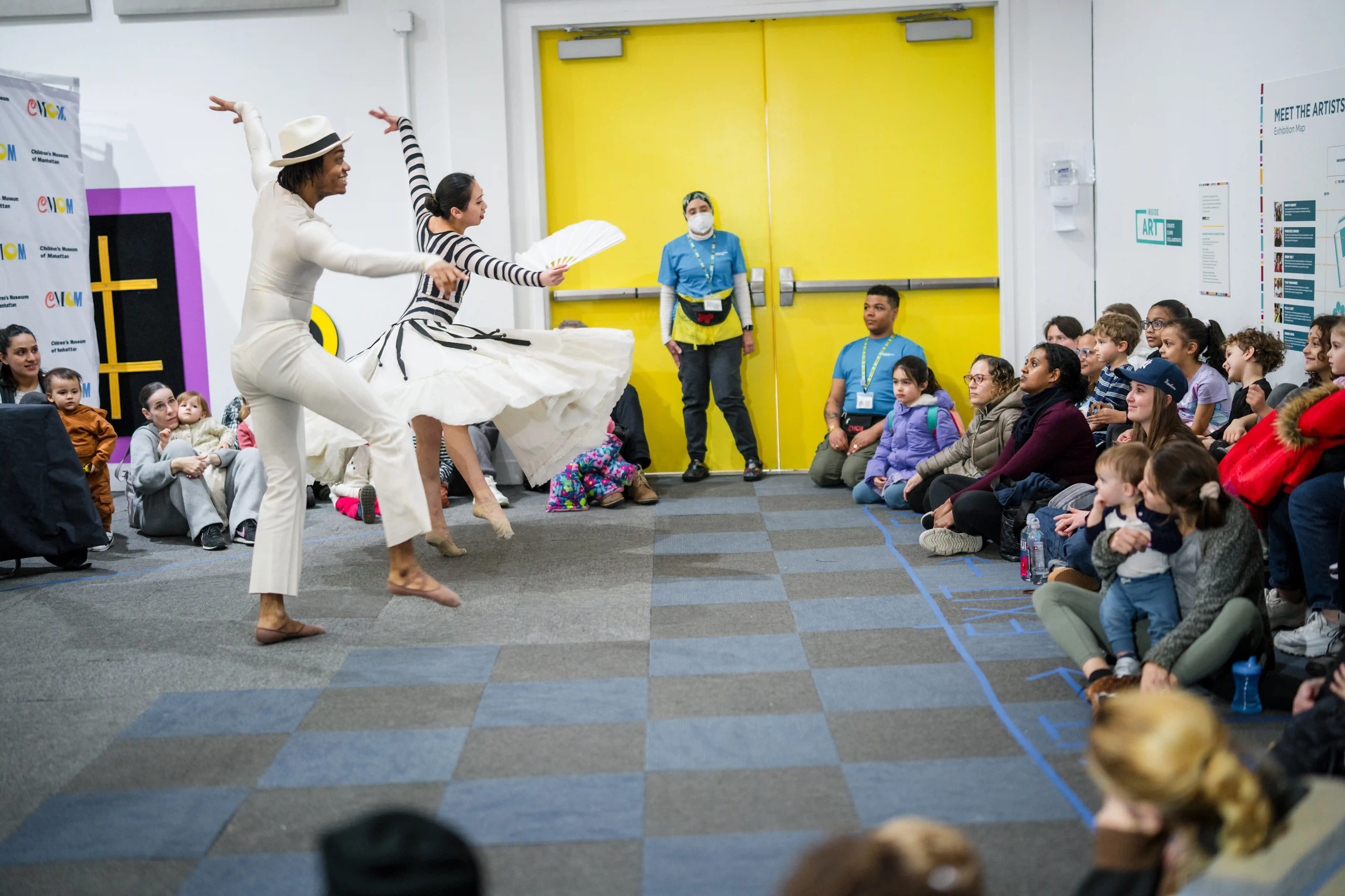 Two dancers perform for a crowd of children and adults. One of the dancers is in a white shirt and white pants with a white hat. The other is in a flowing white skirt and a black and white striped shirt, and holds a white fan. The crowd is seated while watching the dancers. Bright yellow doors are in the background.