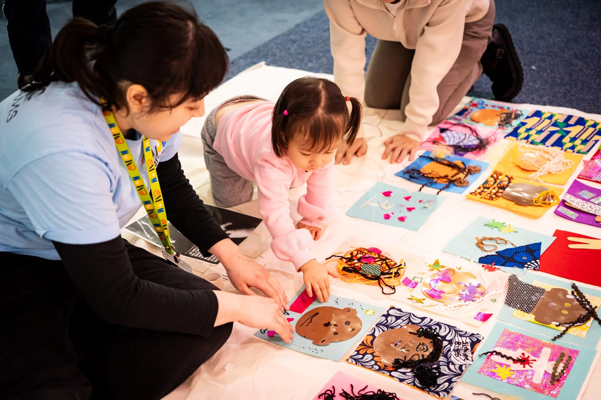 A large cloth is on the floor, with a series of squares that are either different art projects or perhaps meant to be patches for a collective patchwork art piece. A young adult in a Children's Museum of Manhattan Public Programs shirt sits and points out a square to an infant, who is crawling on the floor towards the square. Another adult is partially pictured kneeling by the infant in the background.