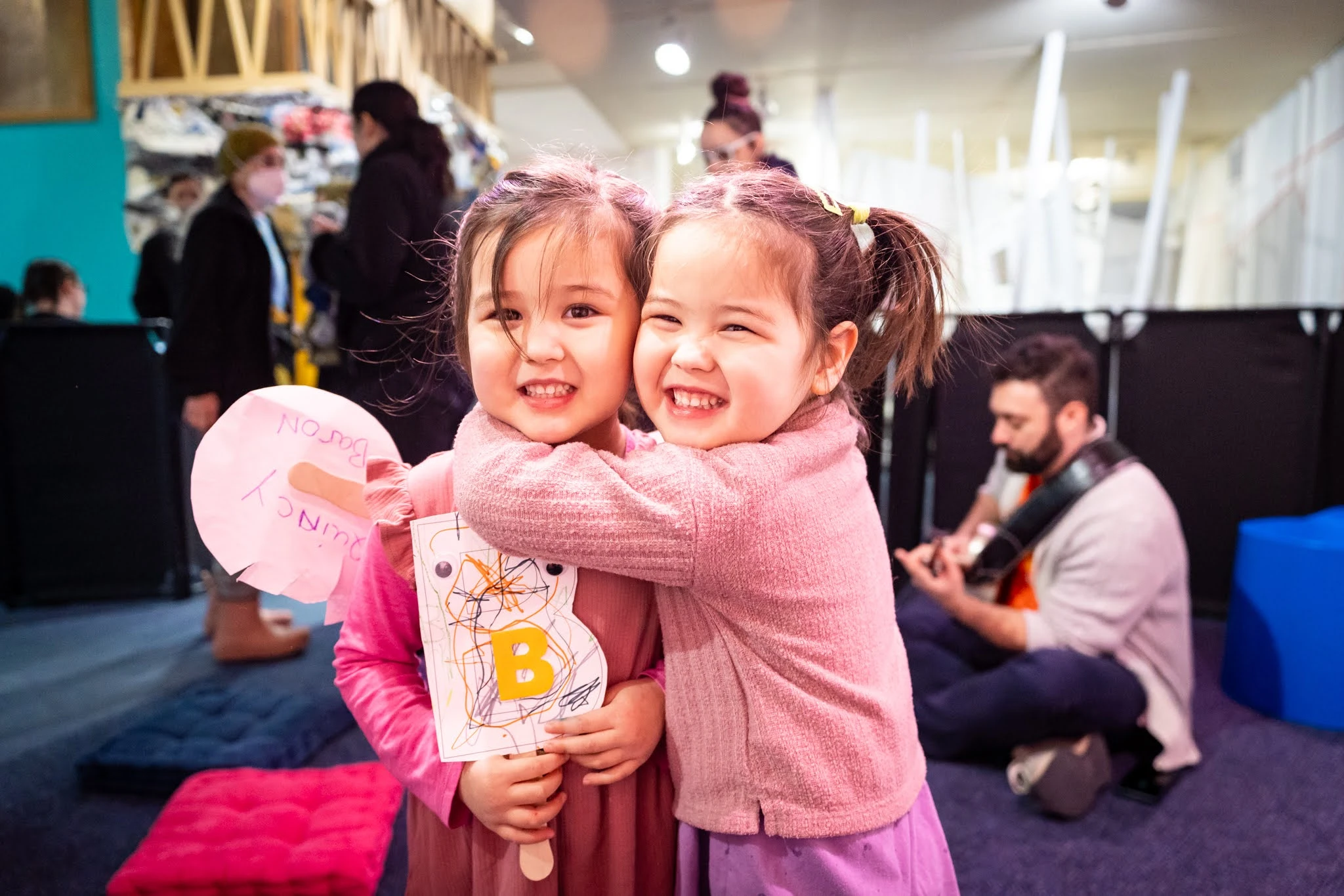 Two children smile for the camera. One embraces the other. Both are holding handmade art on popsicle sticks. In the background are more adults, including one who sits on the floor with a guitar.