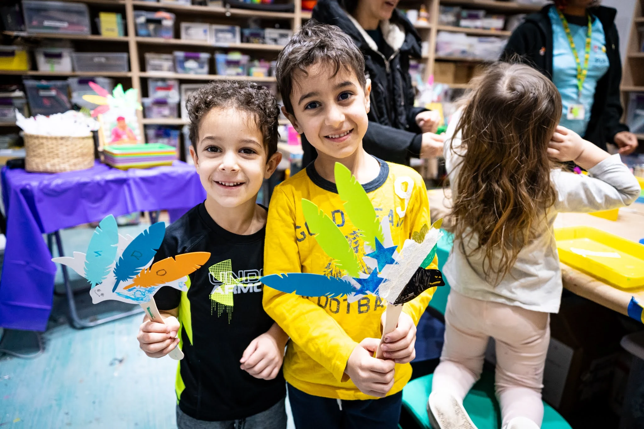 Two children smile for the camera and hold handmade crafts resembling birds. A third child is seen in the background, facing the opposite direction.