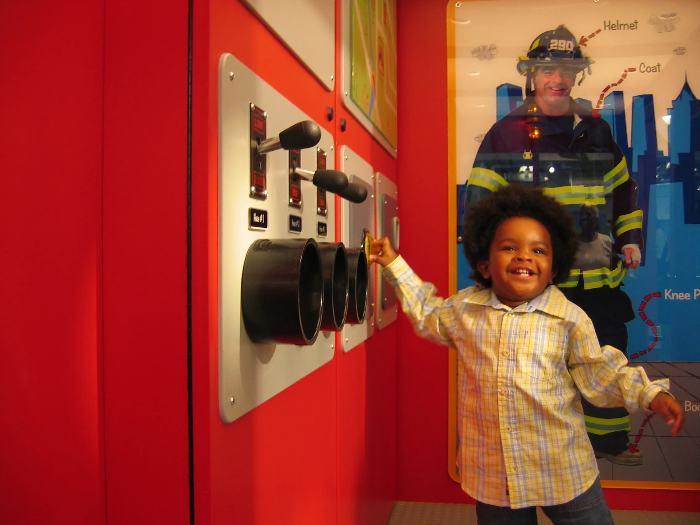 A child smiles at the camera while playing with an exhibit at the Children's Museum of Manhattan. Behind the child, the exhibit includes a photo of an adult fireman in uniform.