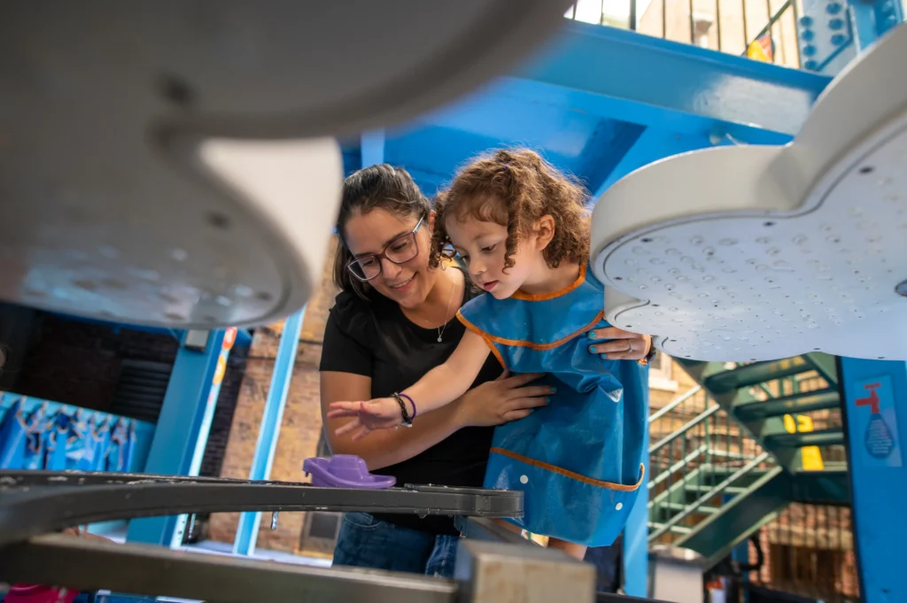 An adult woman holds on to a child, who wears a blue smock. The adult and child are both looking at a water table.