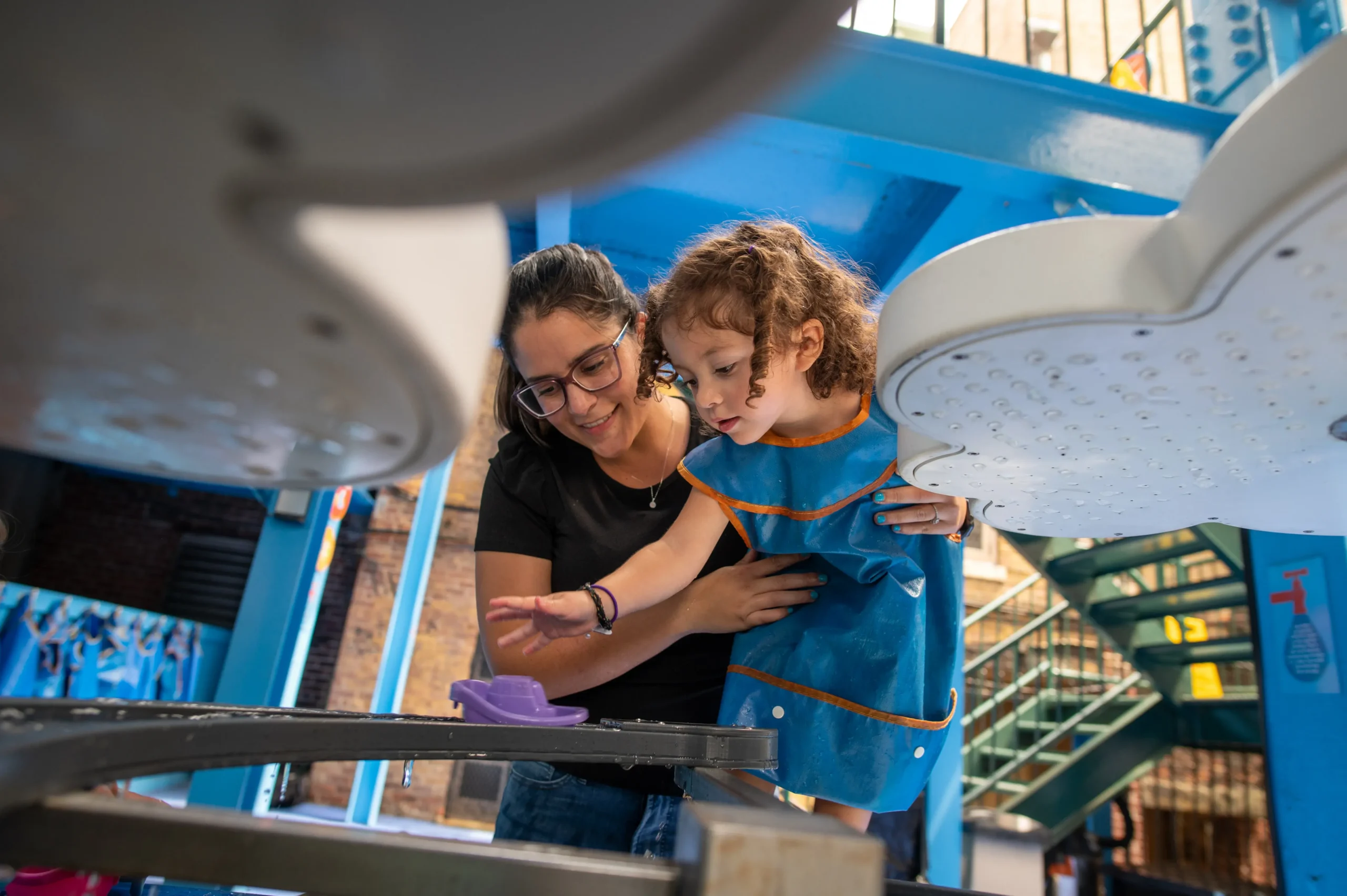 An adult woman holds on to a child, who wears a blue smock. The adult and child are both looking at a water table.