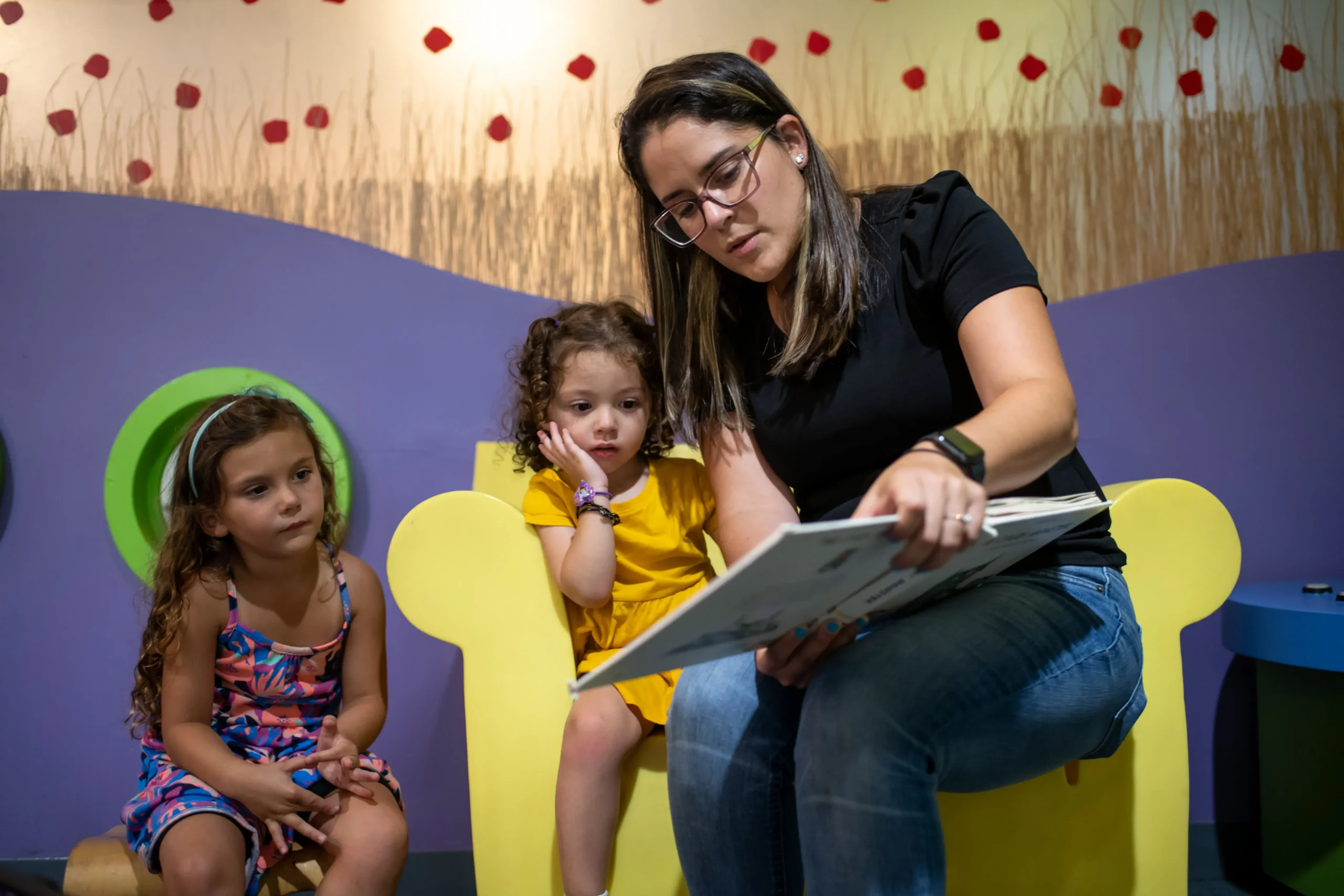 An adult reads to two children. One of the children shares a yellow chair with the adult. All three are looking at the book.