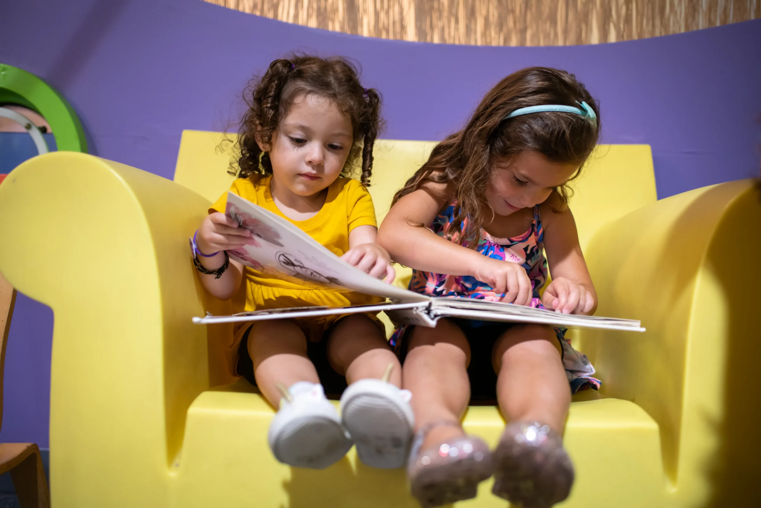 Two children sit in a yellow chair together. They are both looking at the same book, which is laid across their laps.