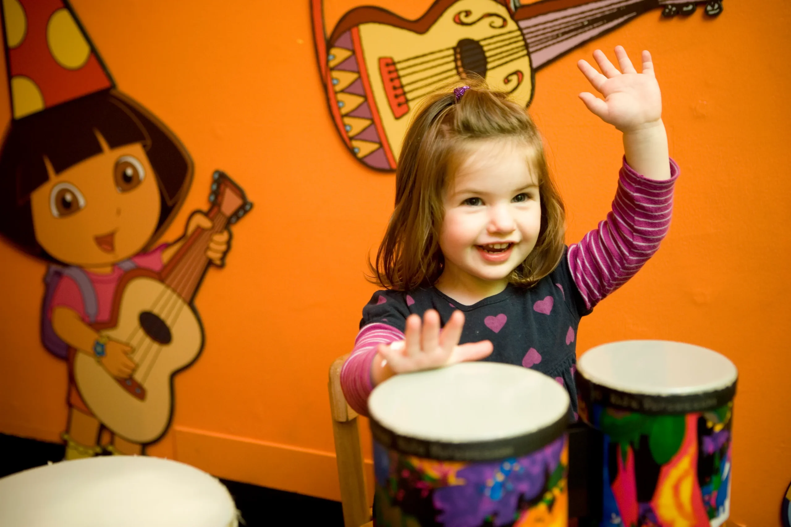 A child sits at bongo drums and has her hands in the air, mid-drum. Behind her is an illustrated version of Dora from the Dora The Explorer cartoon. Dora holds a guitar, and an additional illustrated guitar is included at the top of the frame in the background behind the child.
