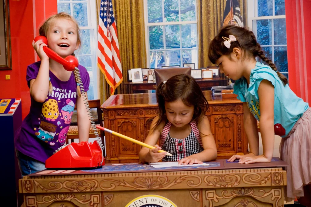 Three small children sit at a toy desk in a toy replica Oval Office. One uses the bright red phone, another uses an oversized pencil and pretends to be writing, and the third watches what the other is writing.