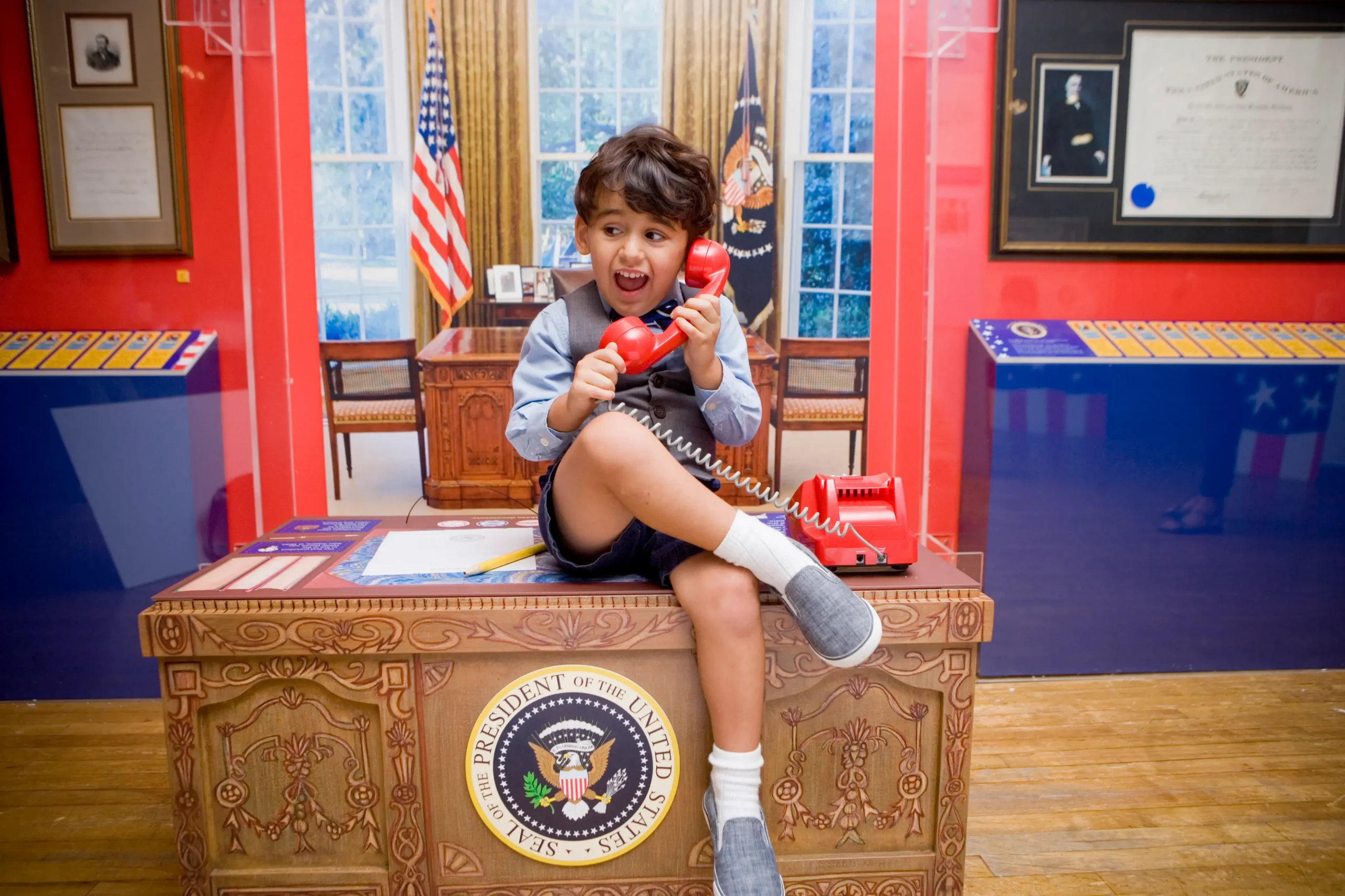A child sits at a toy desk in a toy replica Oval Office with his legs crossed. He pretends to be on the red phone on the desk.