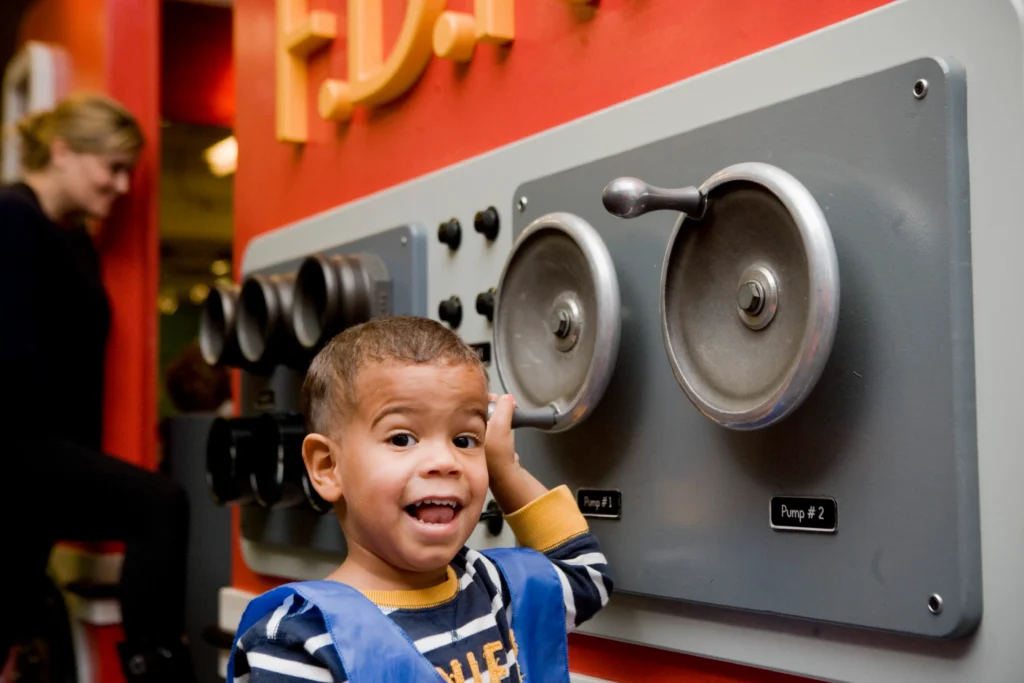A child holds on to a part of a pump that is part of an exhibit at the Children's Museum of Manhattan. He smiles for the camera.