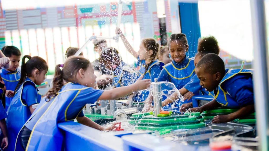 A candid photo of a group of children playing at a water table. All of the children wear matching blue smocks.