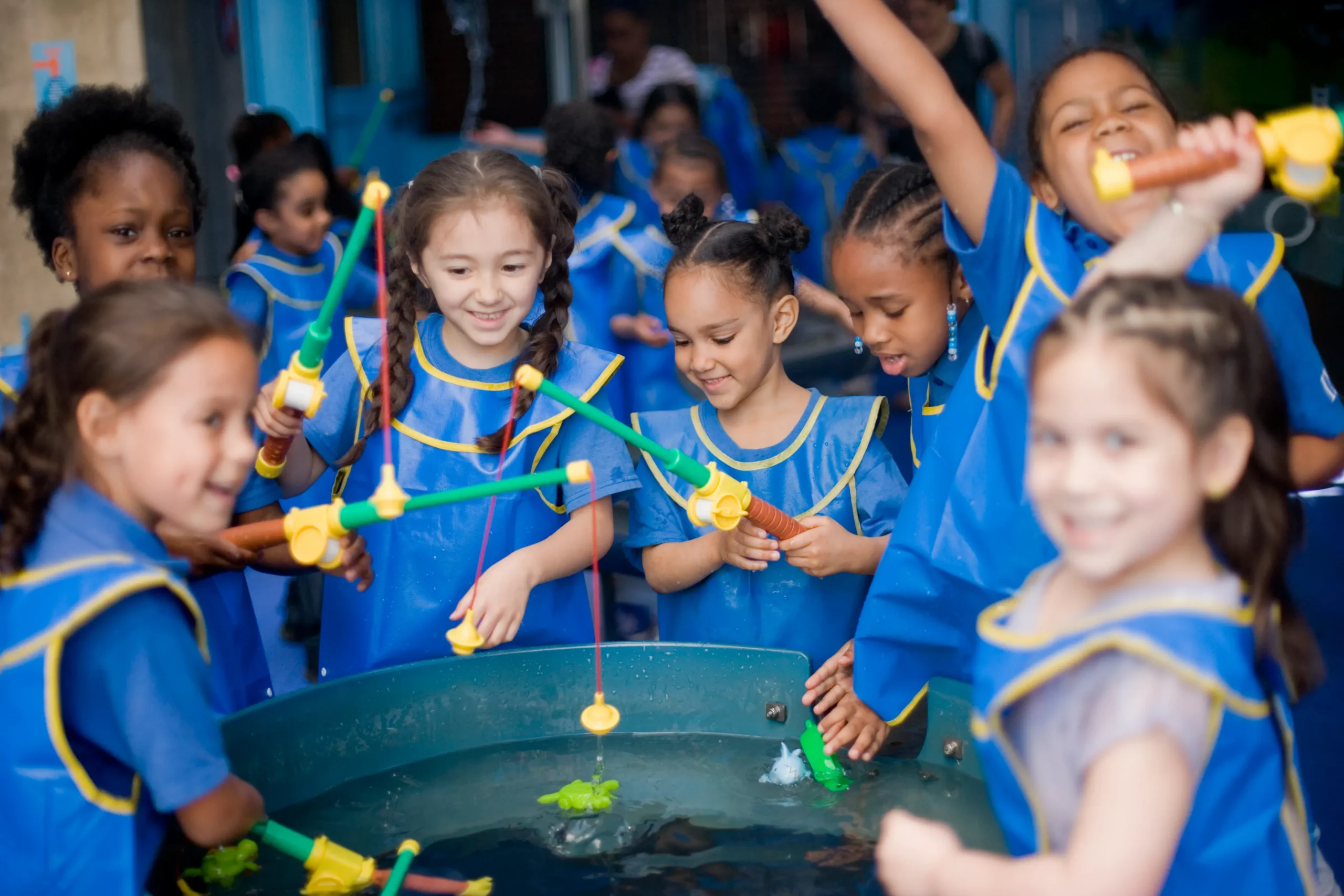 A group of seven children are featured in the front. They all wear matching blue smocks and hold toy fishing poles, playing with them in a water table in front of them. more children are seen in the background.