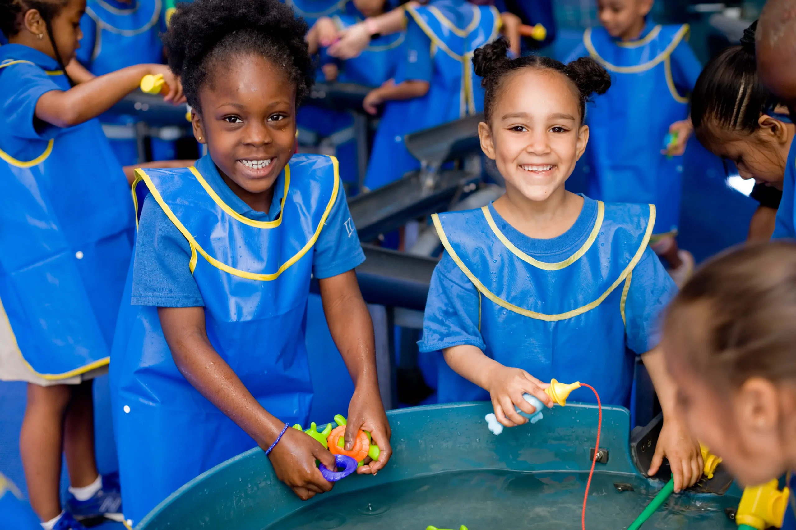 Two children smile at the camera. They are in matching blue smocks and hold water toys. They are standing at a water table. More children in smocks are visible in the background.