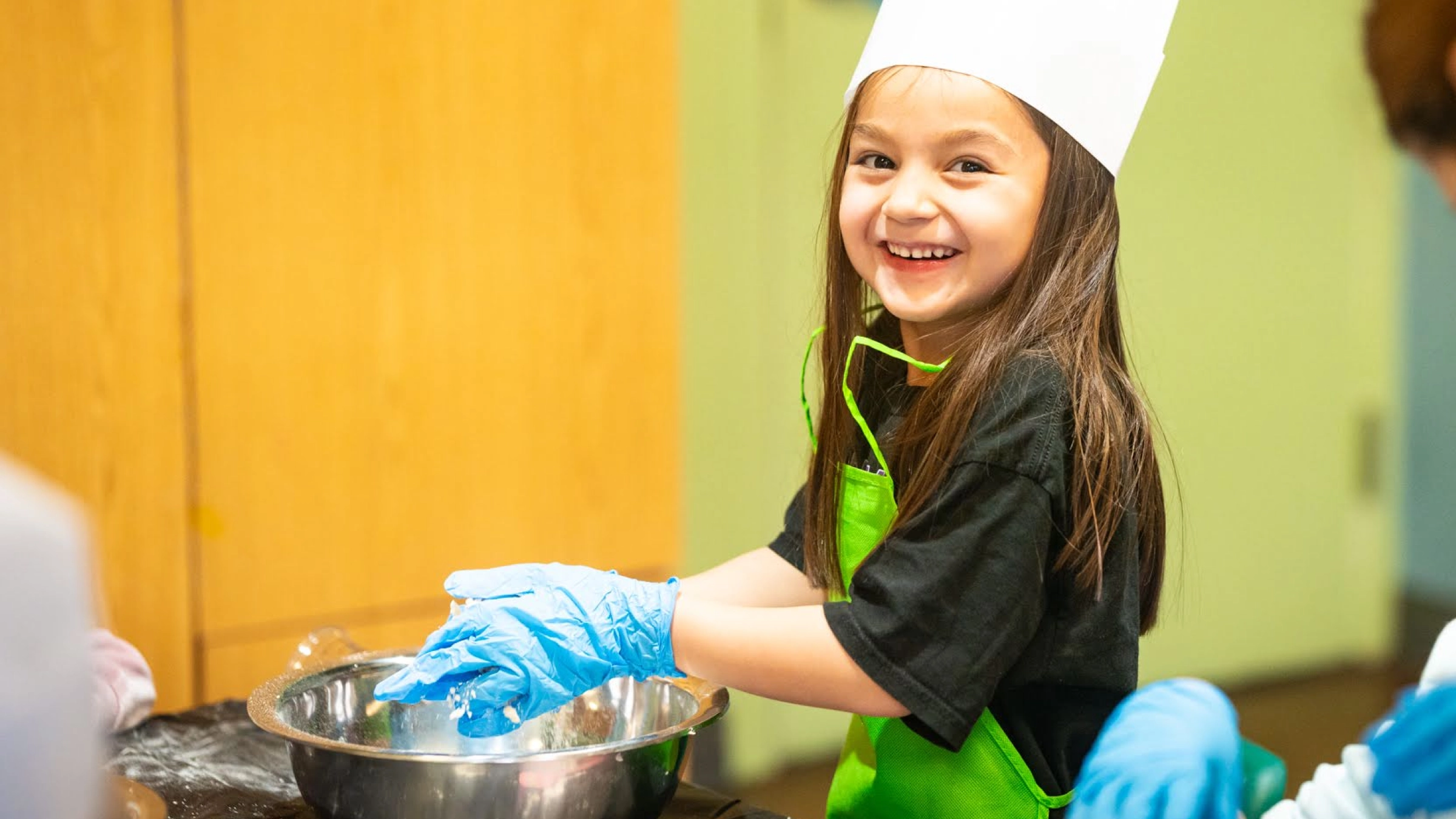 A smiling child in rubber gloves, an apron, and chef's hat, mixing together ingredients in a bowl