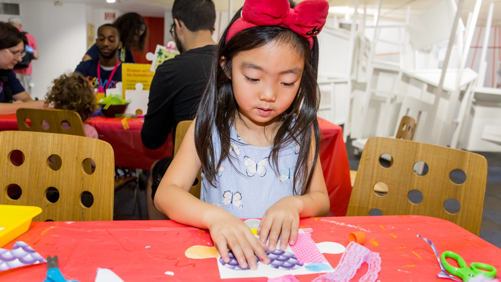 A young child with a large red bow in her hair, assembling crafts together on a table.