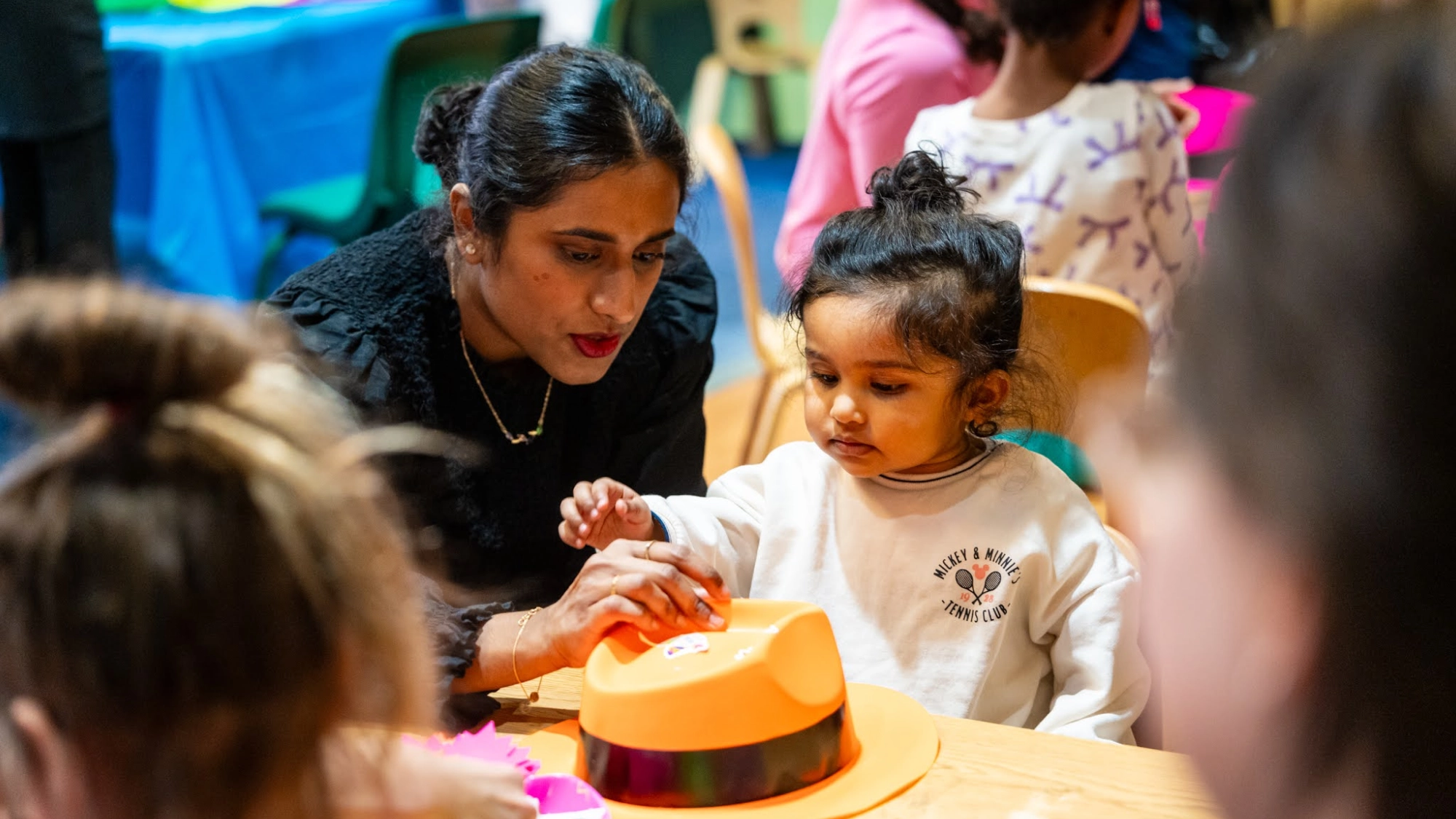 A woman guides a young toddler at Children's Museum of Manhattan, both examining a hat on a table.