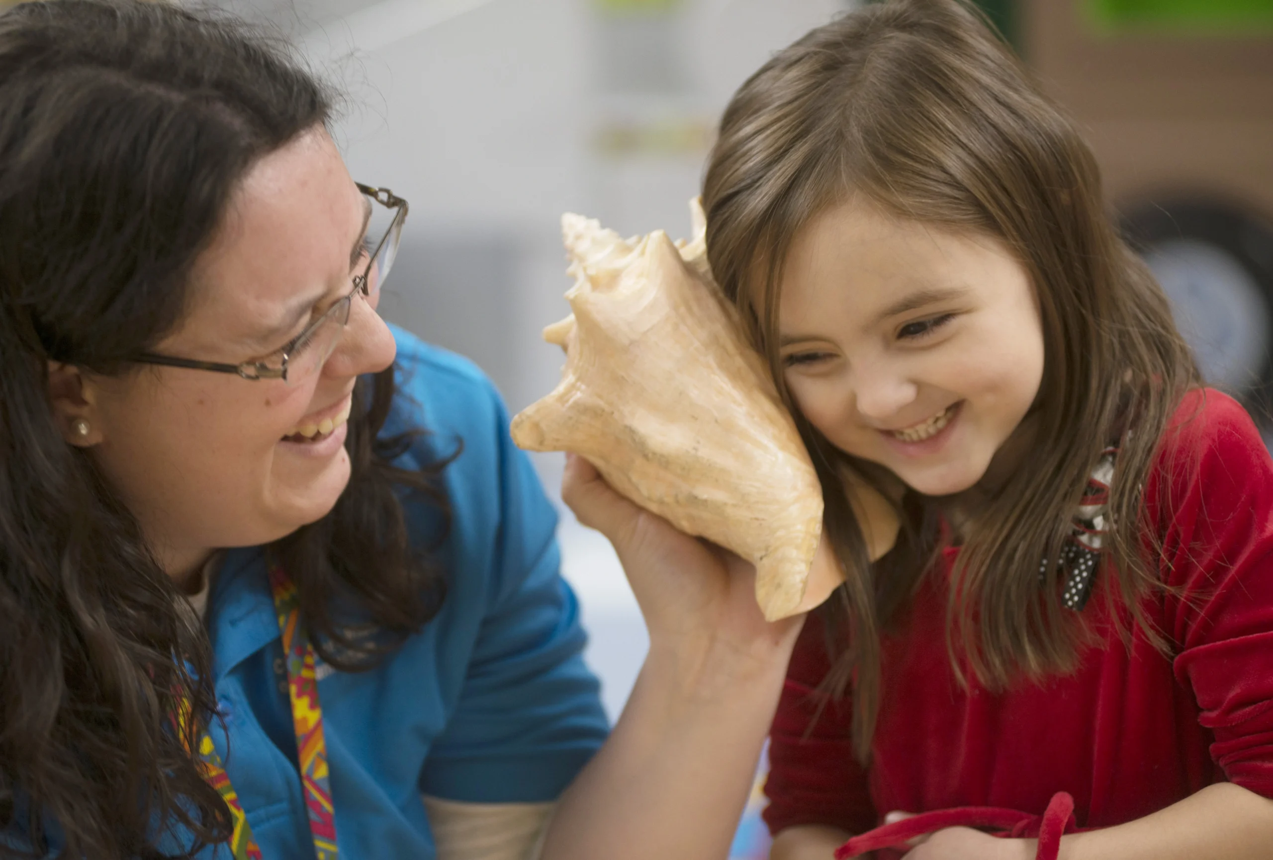 An adult looks at a child and holds a conch shell up to the child's ear. Both the adult and child are smiling.