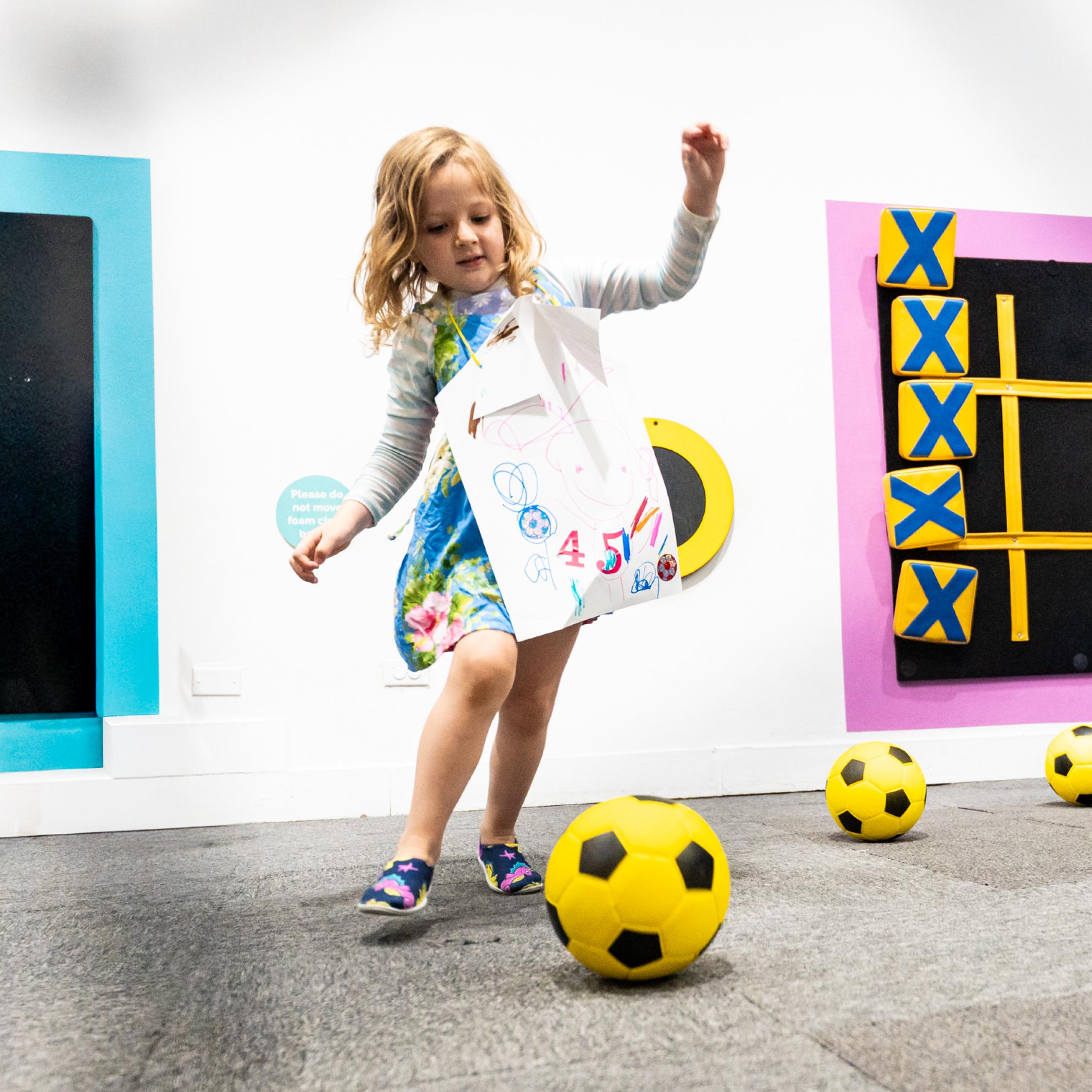 Wearing a handmade paper soccer jersey, a child kicks a soccer ball into a net.