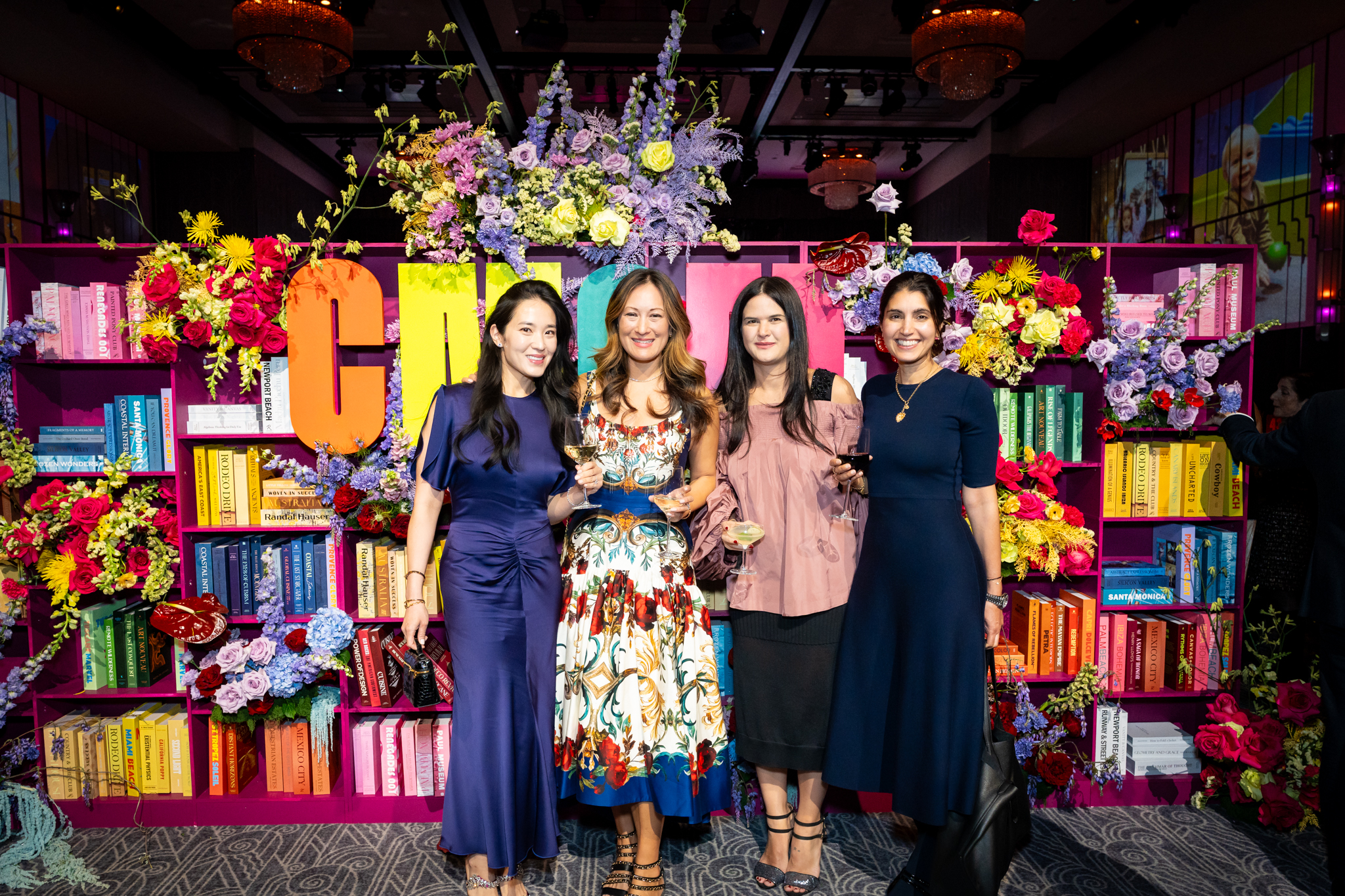Four women in formal dress smile at the camera in front of a colorful bookshelf photo backgrop. Children's Museum Of Manhattan photos by @StewPhotographyNYC
