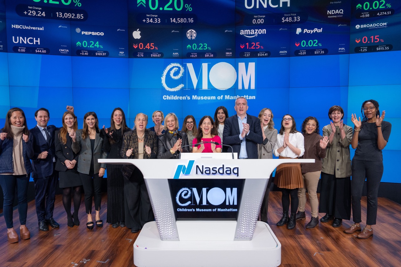 Large group of adults in front of screens with "CMOM" logo at NASDAQ