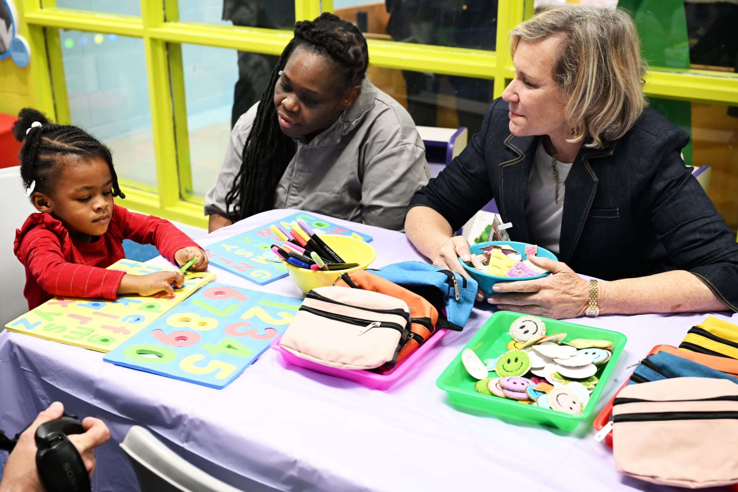 Two adults sit at a table with crafts and supervise a young child