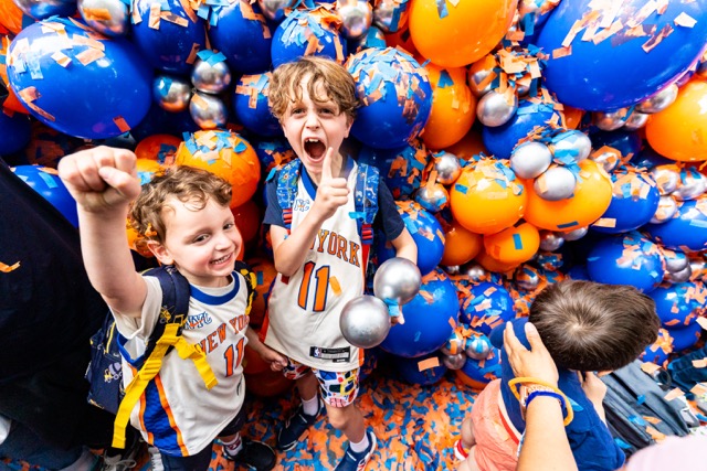 Two children in Knicks jerseys cheer in front of orange and blue balloons with orange and blue tickertape confetti.