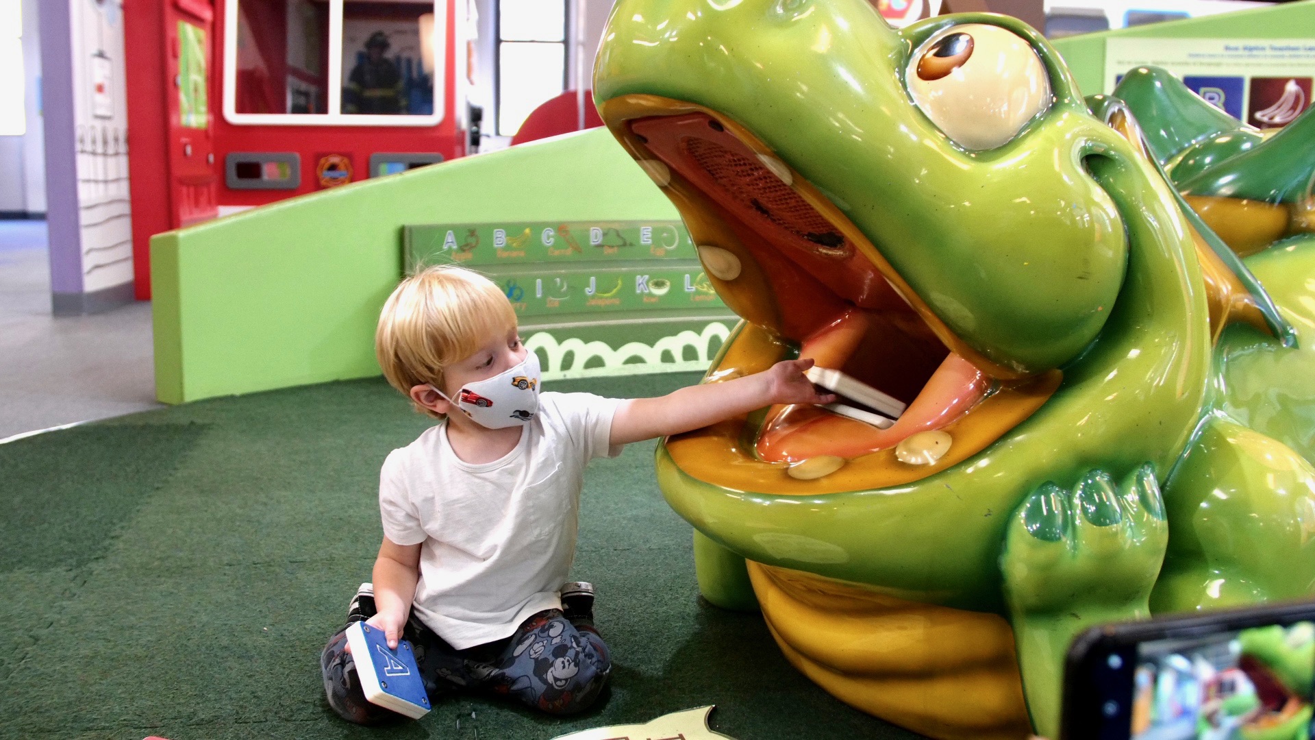 A child in a mask covering his nose and mouth feeds letters to the figure of a dragon at Children's Museum of Manhattan.