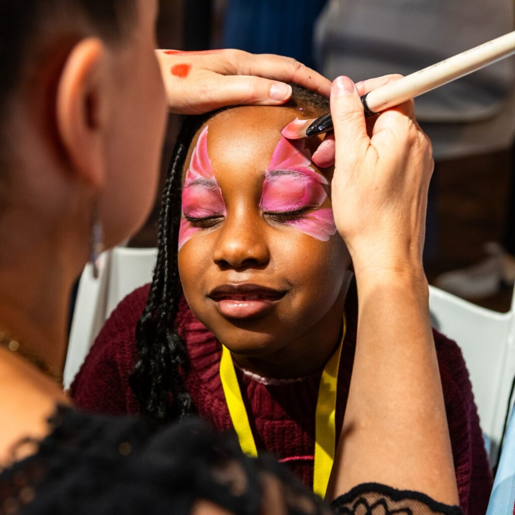 A child smiles with her eyes closed while getting face paint applied by a person who has their back to the camera.