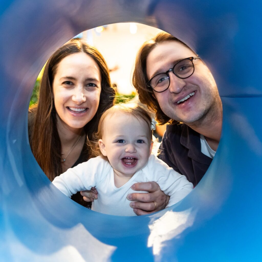Two adults and one baby smile at the camera, as seen through a pipe or large cylinder.