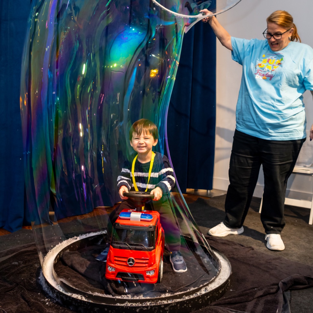A child sits in a firetruck that appears to be in the middle of a giant soap bubble, which an adult behind him is managing.