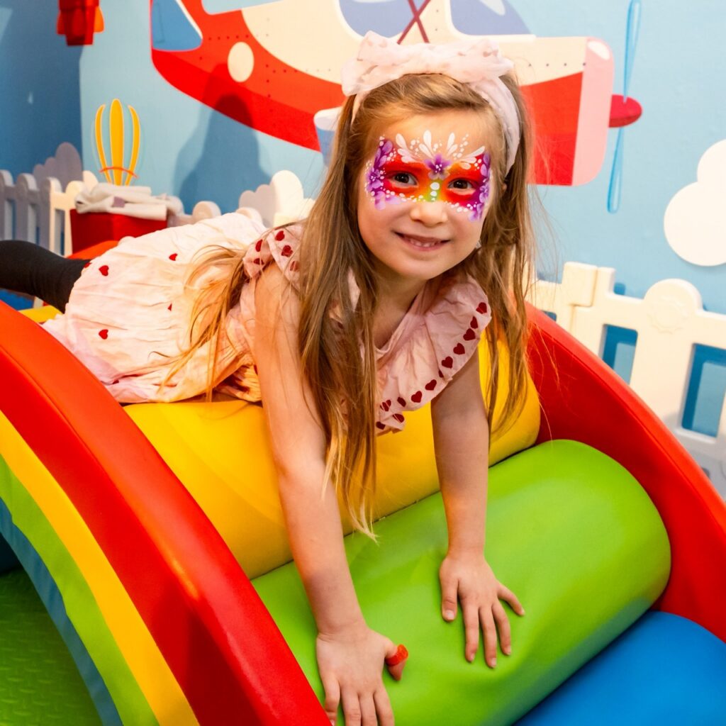 A child in face mask plays on a rainbow slide.