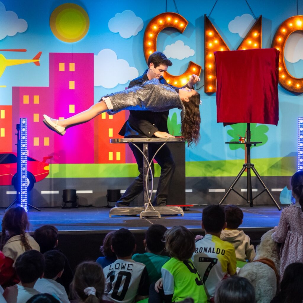 A magician performs a trip in front of an audience, which appears to involve a young child levitating.