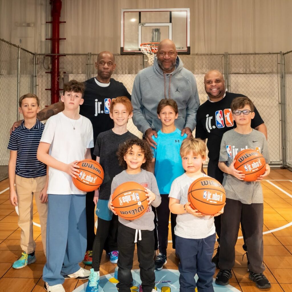 Three adults men with seven young children standing in front of them, four of whom hold basketballs. They all stand in front of a basketball hoop and smile for the camera.