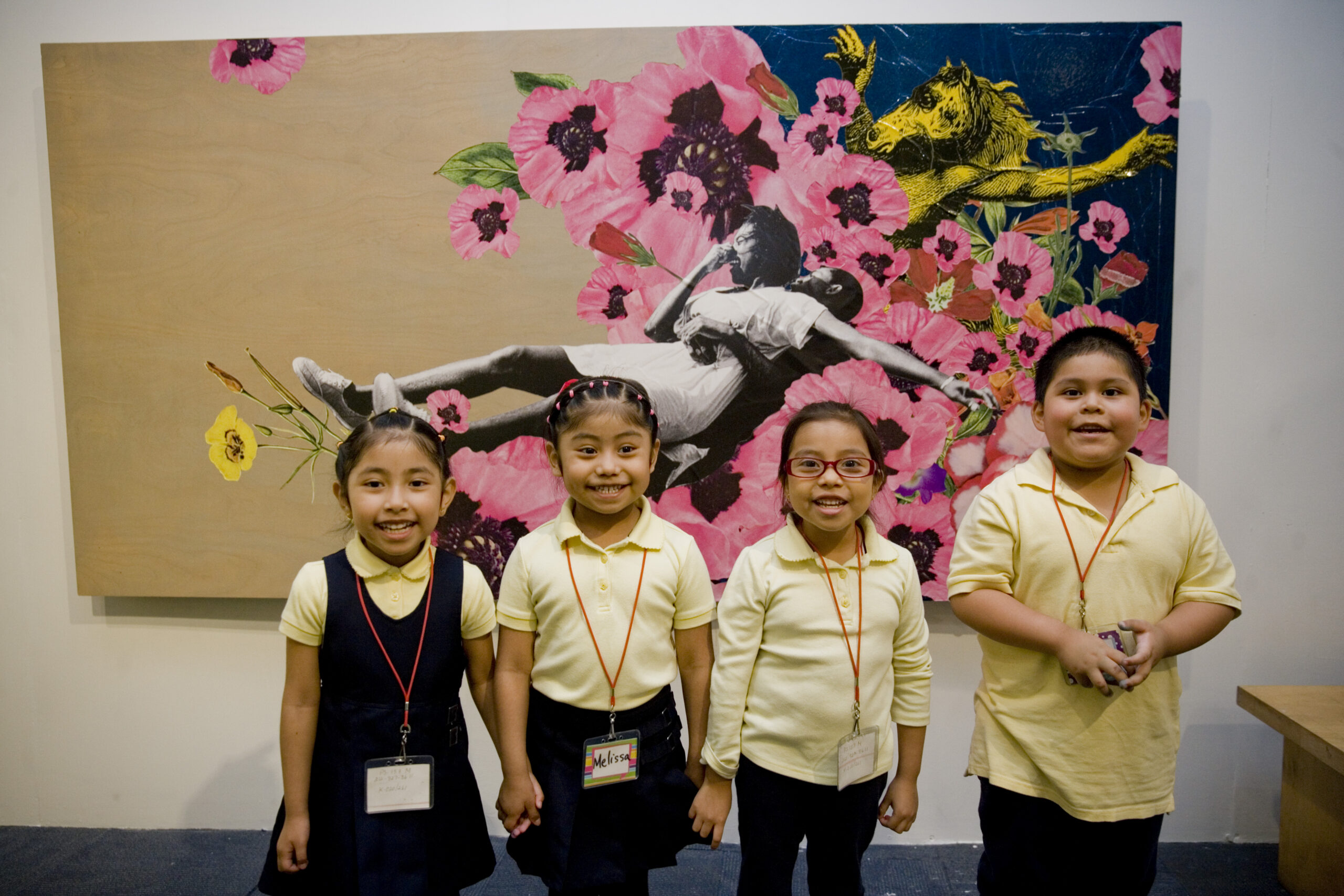 A group of young children in school uniforms smile at the camera in front of a piece of modern art.