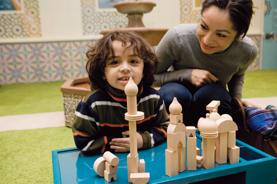 Child smiles at camera while adult watches child. They are both seated on the floor at a toy block version of a mosque.