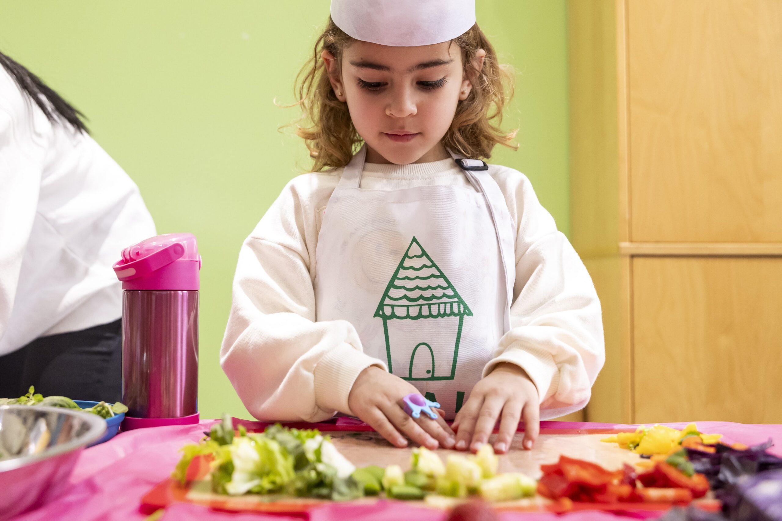 A child in a smock and paper hat at a table with prepped and chopped fruits and veggies in front of her.