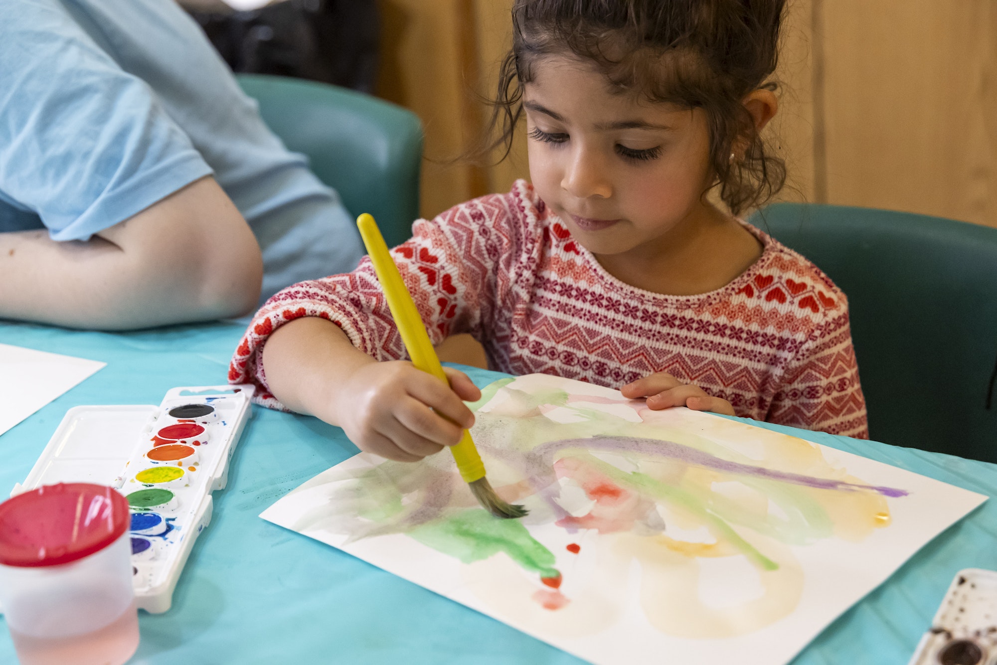 Child paints with simple children's watercolor set on a piece of paper in front of her while she sits at a table.