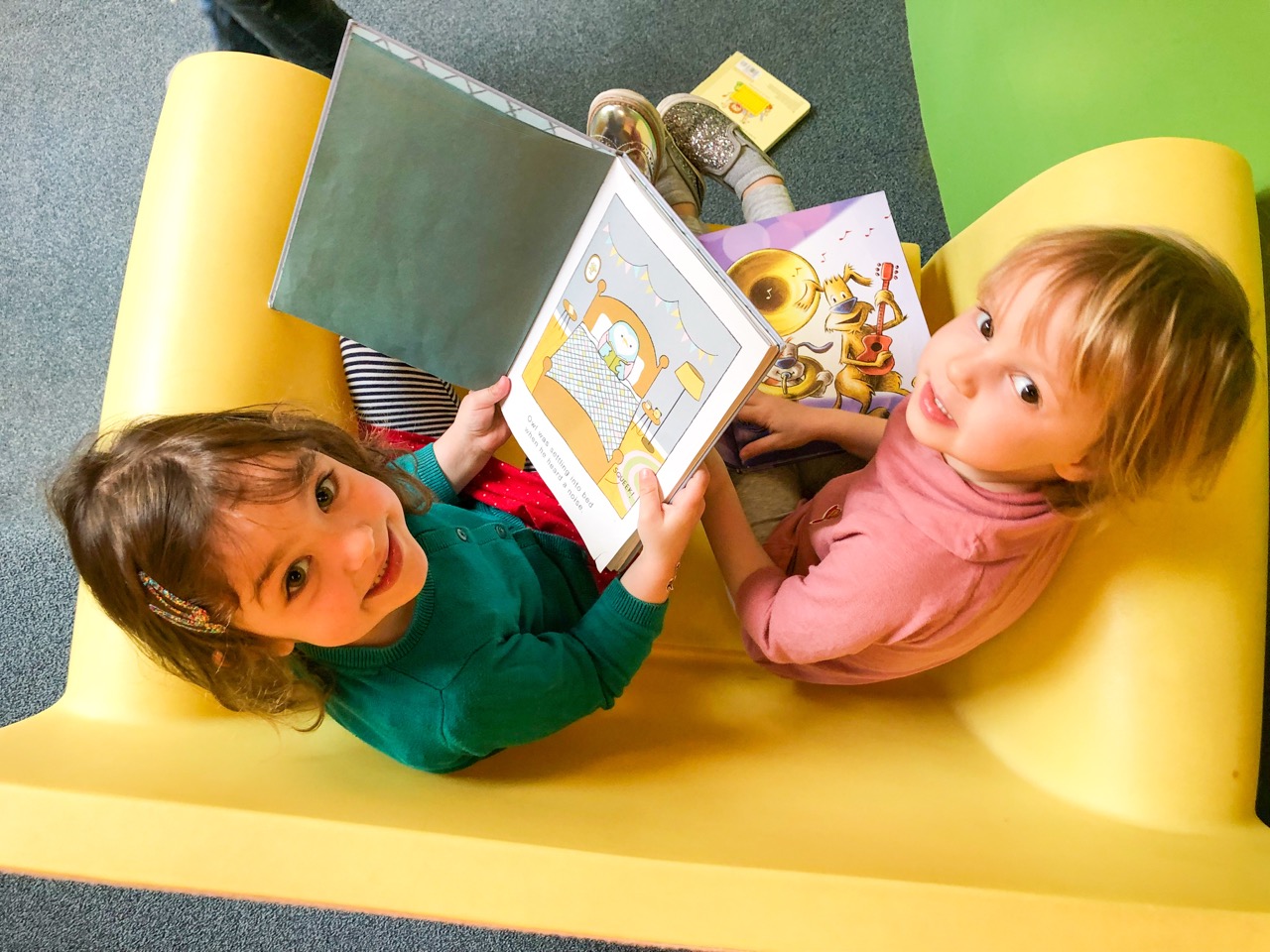 Two small children sit in a chair with books open in their laps. They both look up and smile at the camera.