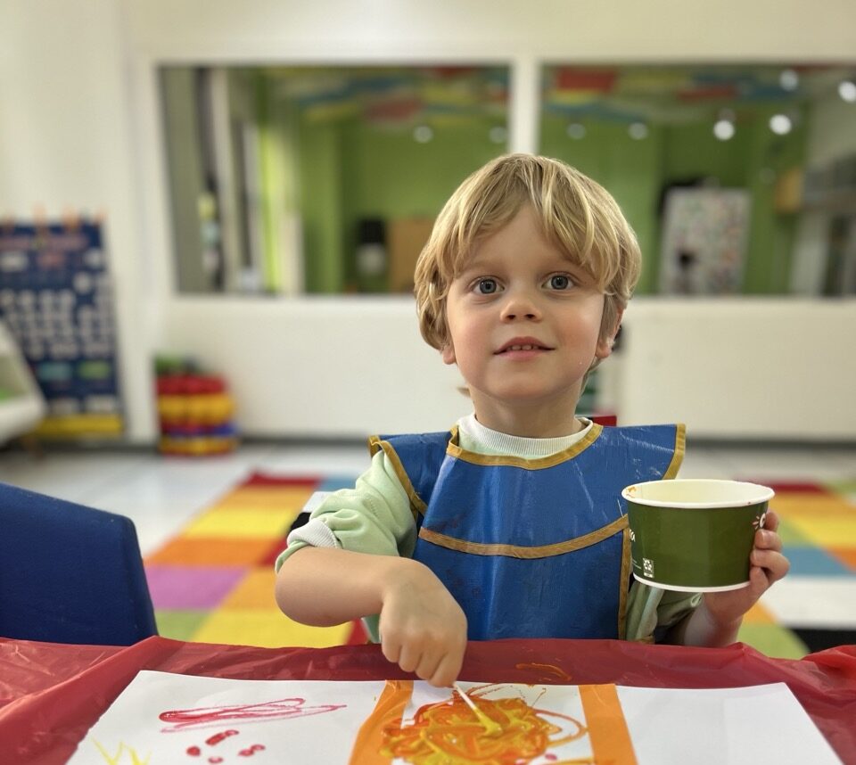 A child looks at the camera while he paints on a piece of paper in front of him.