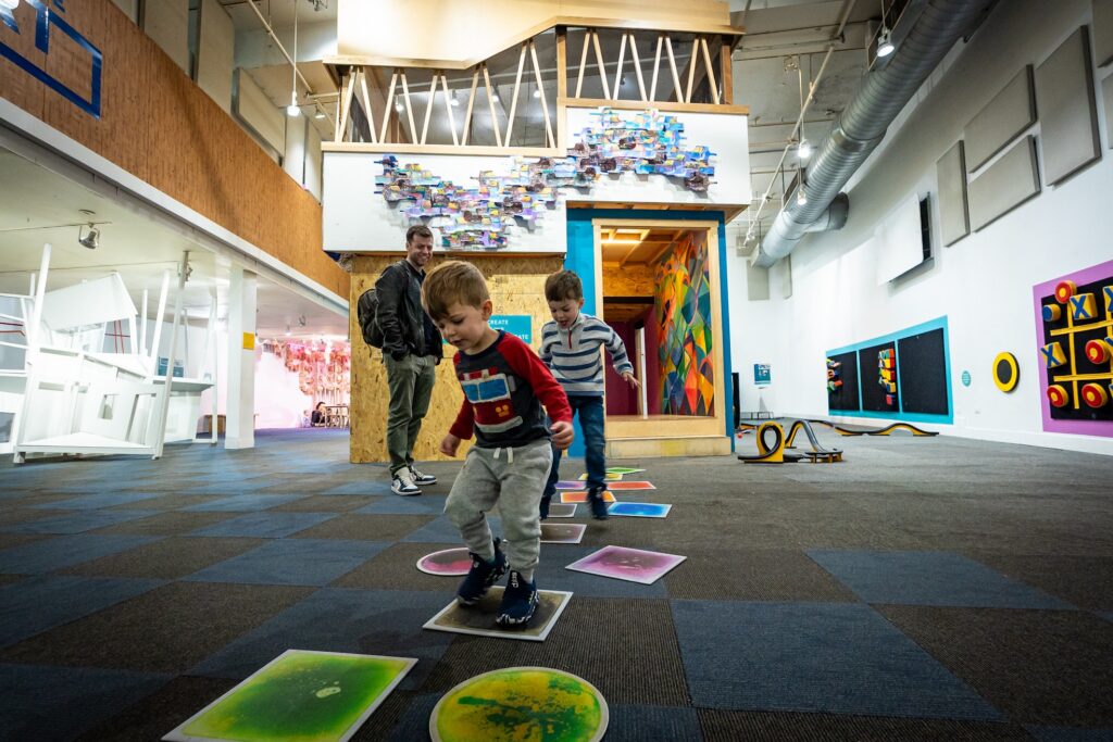Two small children playing on toy floor tiles in front of an art exhibit. An adult wearing a backpack looks on and smiles.