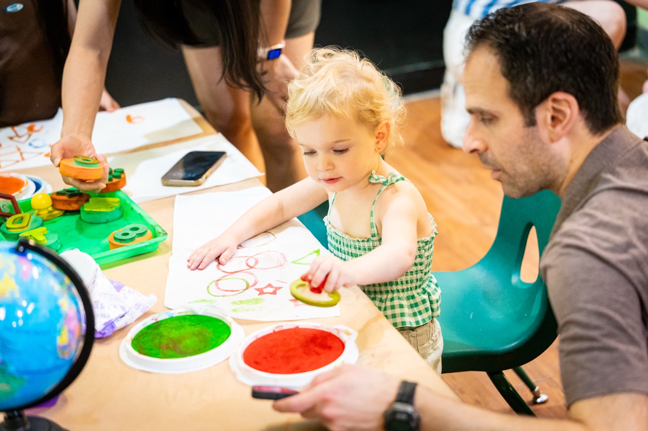 A young child at a craft table painting while an adult supervises